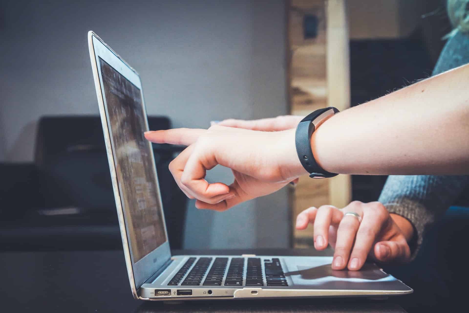 Two hands are interacting with a laptop on a lead generation website. One hand is pointing at the screen, while the other is typing on the keyboard. The person pointing is wearing a black wristband. There are blurred, indistinct objects and furniture in the background.