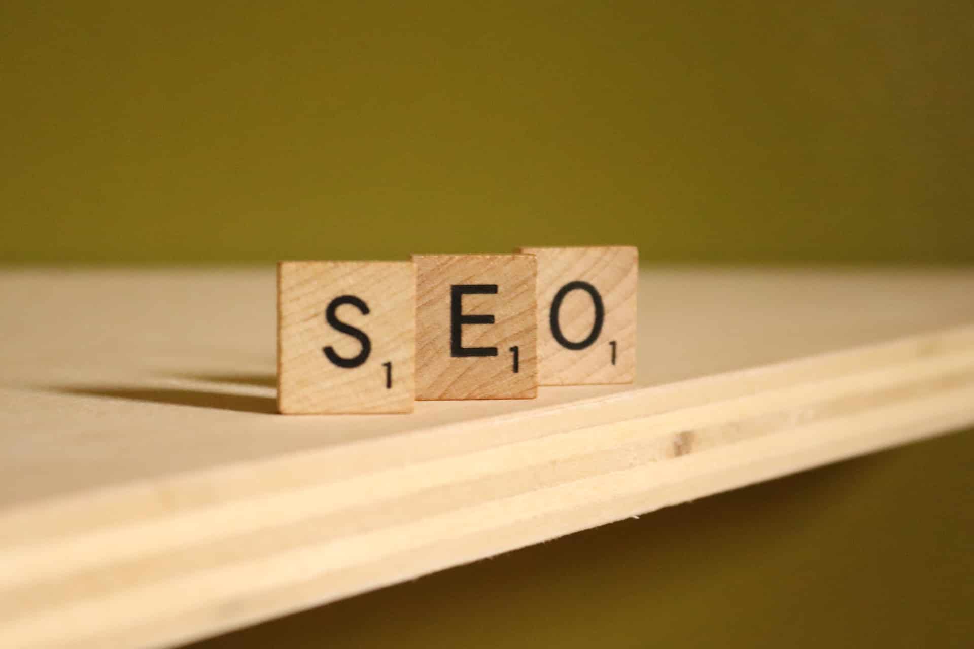 Three wooden Scrabble tiles placed on a wooden surface spell out "SEO" against a plain olive green background. Each tile also displays the number 1.